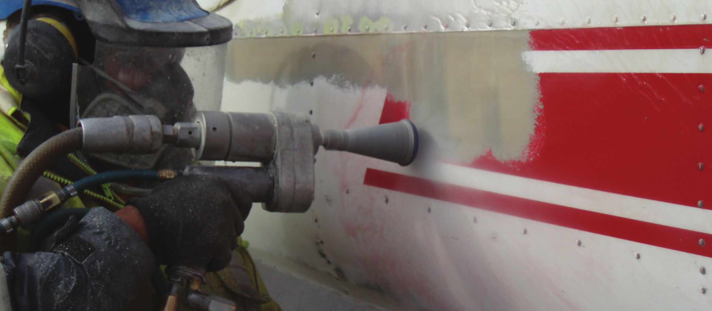 An operator in a hood and hi-vis holding a lance that strips red and white paint from an aircraft fuselage