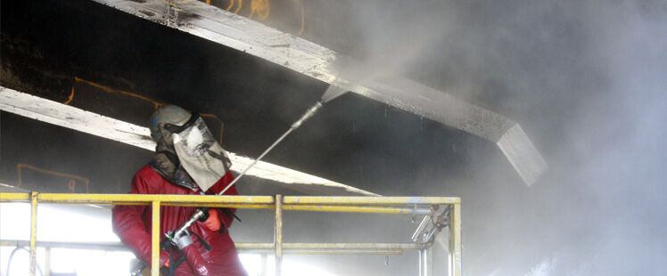 An operator in a red protective suit on a bridge deck behind a wide plume of spray