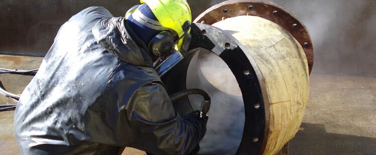 Two operators in protective gear blasting scale from a turbine casing