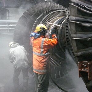 Two operators in hi-vis blasting a large turbine rotor with spray drifting across the frame